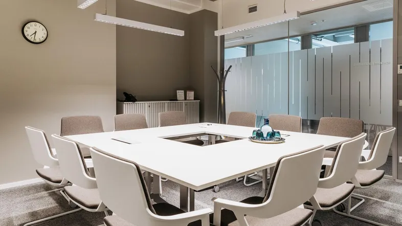 Moderner Konferenzraum mit großem quadratischem Tisch, bequemen Stühlen und Glaswänden für eine professionelle Arbeitsumgebung.