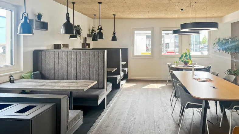 Moderner Gemeinschaftsbereich mit Sitznischen, langen Tischen und Holzboden für entspanntes Arbeiten oder Pausen.