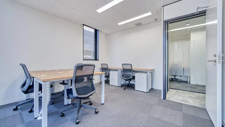 Modernes Büro mit mehreren Arbeitsplätzen, Teppichboden und einer Glastür zu einem weiteren Raum.