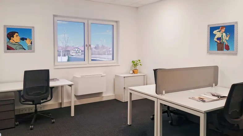 Heller Büroraum mit zwei modernen Arbeitsplätzen, Teppichboden und einem Fenster mit Blick ins Grüne.