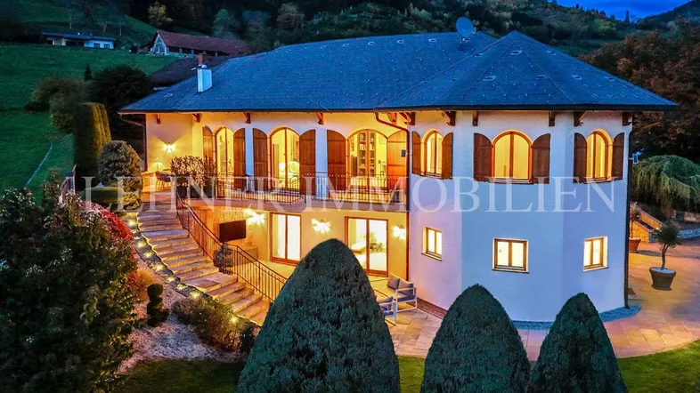 Elegante Villa bei Dämmerung mit beleuchteter Fassade und gepflegtem Garten in bergiger Landschaft.