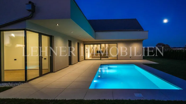 Modernes Haus bei Nacht mit beleuchtetem Pool und großzügiger Terrasse, umgeben von Rasenfläche.