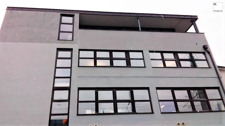 Moderne Gebäudefassade mit großen Fenstern und einem Balkon im obersten Stockwerk.