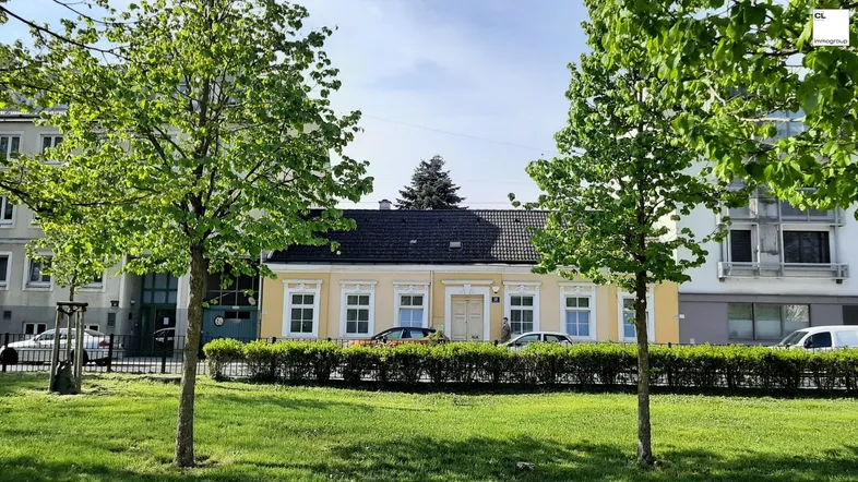 Gelbe Fassade eines Hauses mit schwarzem Dach, umgeben von Bäumen und Grünflächen.