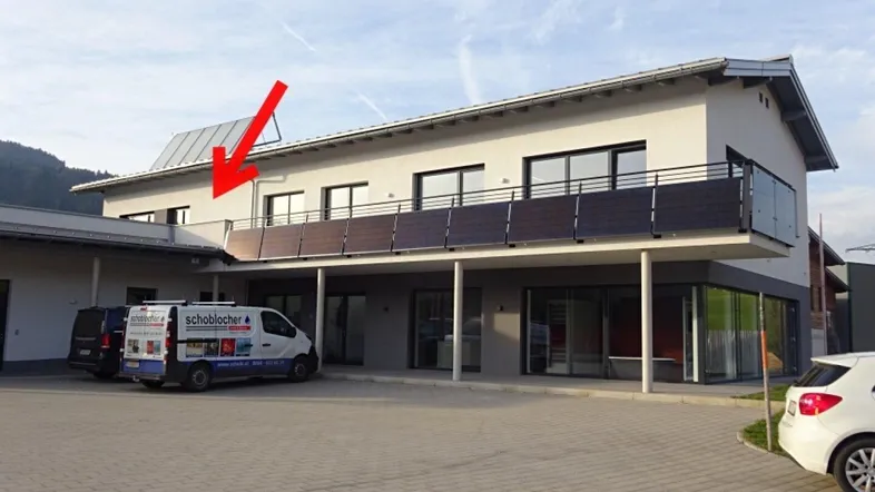 Modernes Geschäftsgebäude mit Balkon im Obergeschoss und Parkplätzen davor unter einem klaren Himmel.