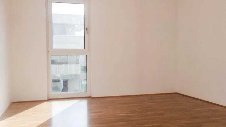 Helles, leeres Zimmer mit Holzboden und großem Fenster, das viel Tageslicht hereinlässt.