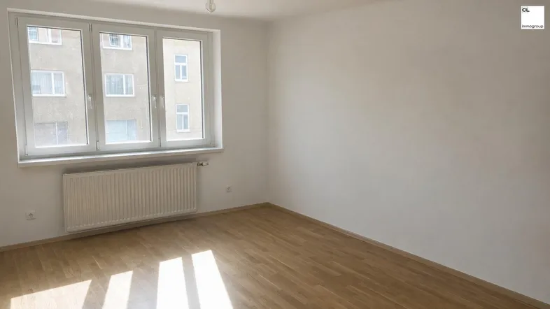 Helles Zimmer mit Parkettboden und Blick auf die umliegenden Gebäude, ausgestattet mit einem Heizkörper unter dem Fenster.