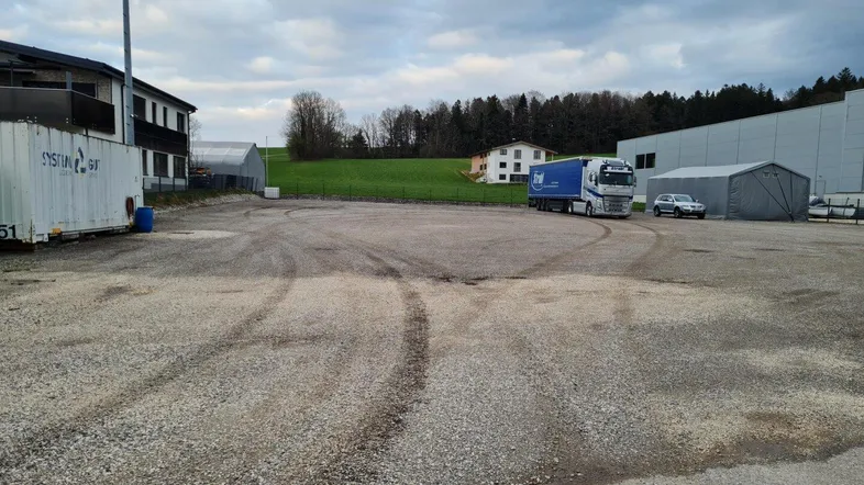 Großer Schotterparkplatz mit Lastwagen und angrenzenden Gebäuden unter bewölktem Himmel.