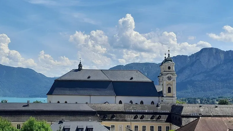 Panoramablick auf ein historisches Gebäude mit See und majestätischen Bergen im Hintergrund unter blauem Himmel.