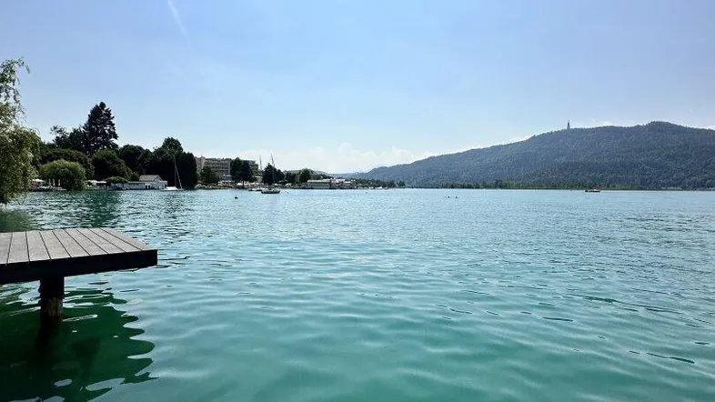 Seeufer mit Holzsteg und malerischem Blick auf die umliegende Berglandschaft unter blauem Himmel.