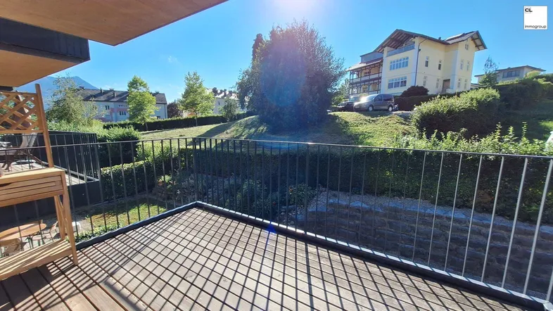 Heller Balkon mit Holzboden und Blick auf grüne Hügel und umliegende Gebäude.