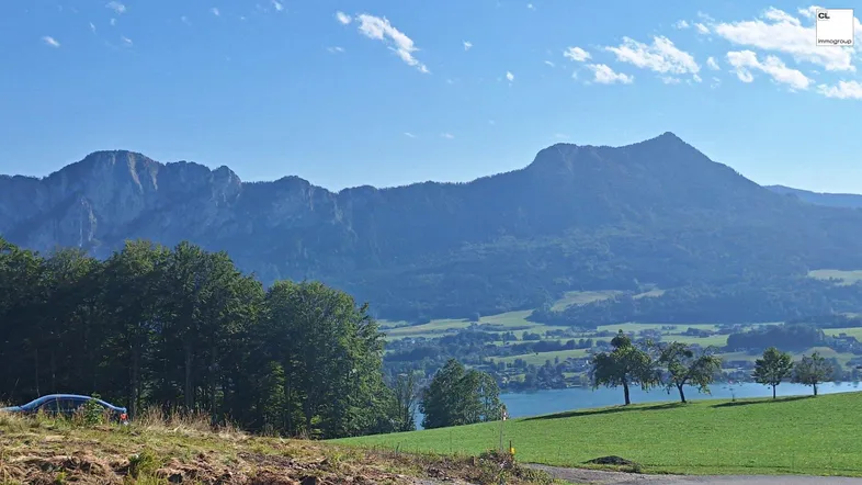 Panoramablick auf majestätische Berge, einen ruhigen See und grüne Hügellandschaft unter blauem Himmel.