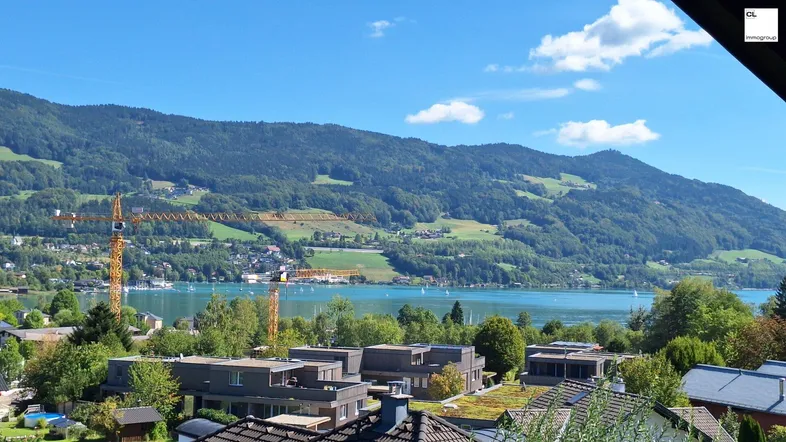 Panoramablick auf den See und die umliegenden Berge mit einigen Gebäuden im Vordergrund unter blauem Himmel.