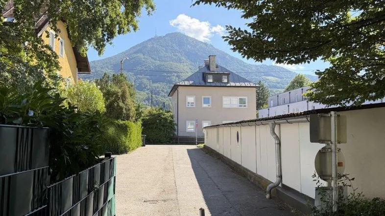 Ein asphaltierter Weg führt zu einem Gebäude mit einem majestätischen Berg im Hintergrund, umgeben von üppigem Grün.