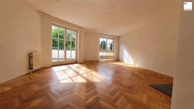 Helles Wohnzimmer mit Fischgrätparkettboden, großen Fenstern und Zugang zu einem Balkon.