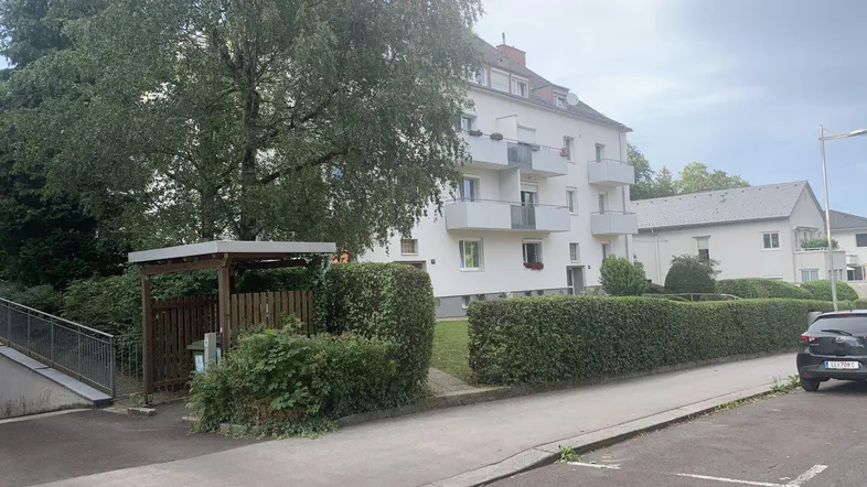 Mehrfamilienhaus mit weißer Fassade, Balkonen und gepflegter Hecke in einer grünen Umgebung.