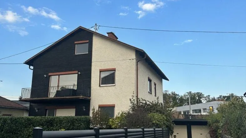 Zweigeteilte Hausfassade mit schwarzem und hellem Putz, umgeben von einem gepflegten Zaun und Vorgarten.