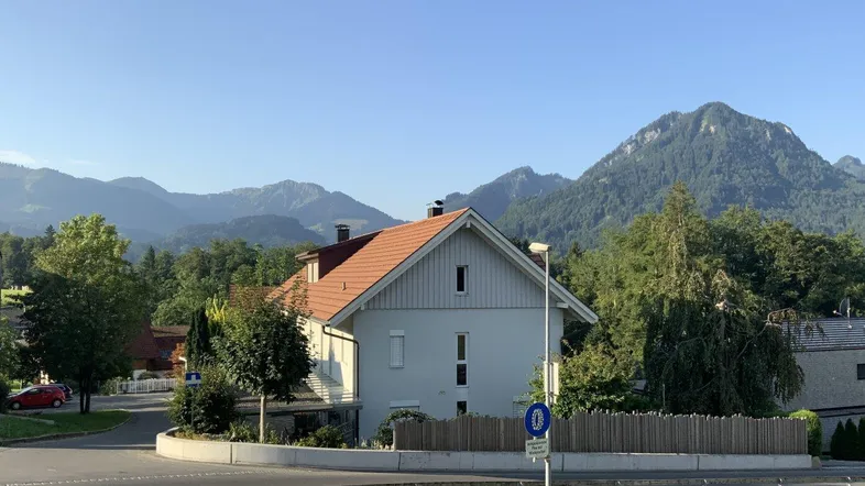 Haus mit rotem Dach und heller Fassade, umgeben von Bäumen und Bergen in ländlicher Umgebung.