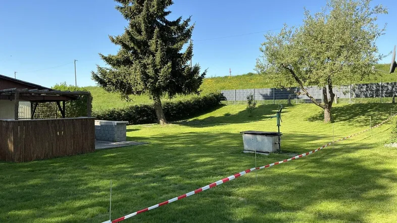 Weitläufiger Gartenbereich mit altem Baumbestand, einem Brunnen und viel Grünfläche unter blauem Himmel.