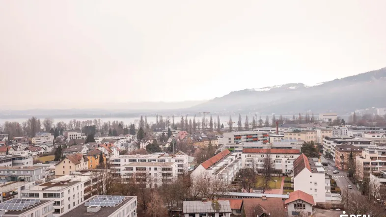 Panoramablick über die Stadt mit See und Bergen im Hintergrund an einem bewölkten Tag.