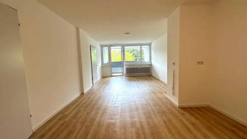 Geräumiges, leeres Wohnzimmer mit hellem Laminatboden und großen Fenstern, die viel Tageslicht hereinlassen.