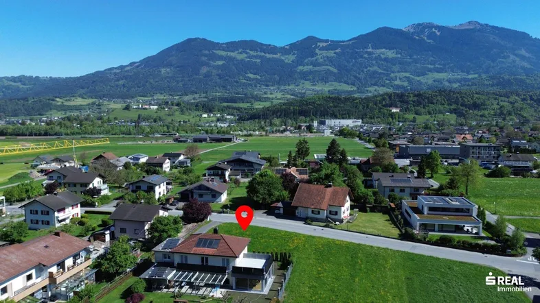 Luftaufnahme einer Wohngegend mit markiertem Grundstück und beeindruckendem Bergpanorama im Hintergrund.