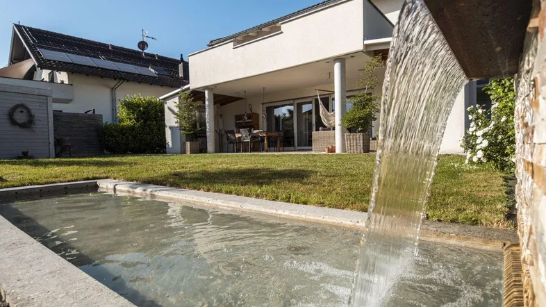 Moderner Garten mit Wasserspiel, Rasenfläche und überdachter Terrasse am Haus.