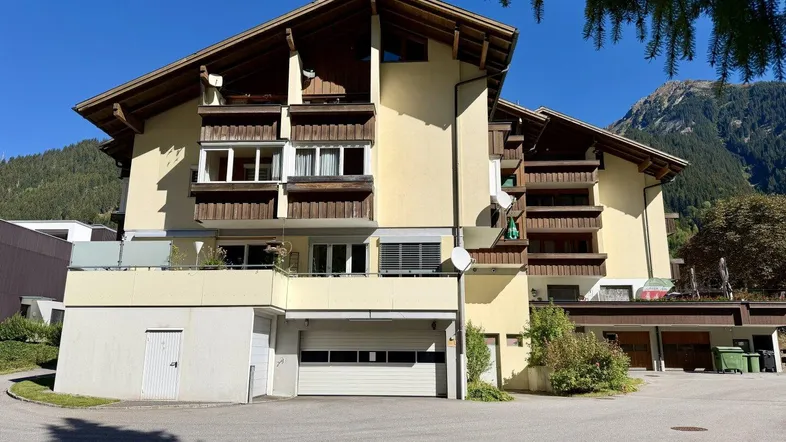 Mehrstöckiges Gebäude mit traditioneller Architektur, Balkonen und integrierter Garage in alpiner Umgebung.