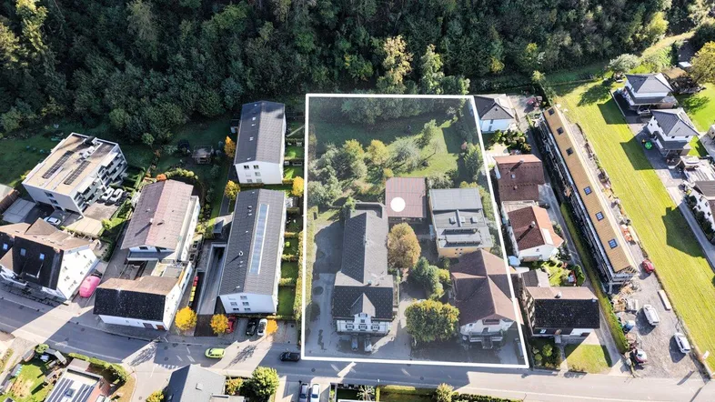 Luftaufnahme eines Grundstücks mit umliegender Bebauung und Wald im Hintergrund.
