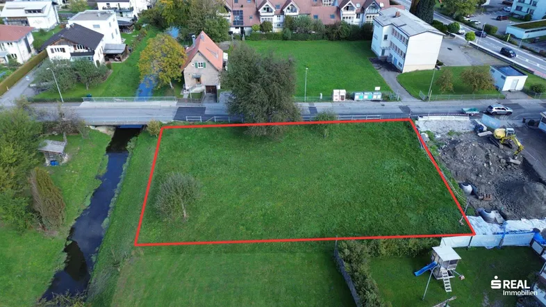 Luftaufnahme eines grünen Baugrundstücks mit roter Umrandung, angrenzendem Bach und umliegenden Häusern.