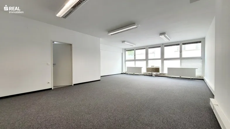 Geräumiges, leeres Büro mit Teppichboden und großen Fenstern, die viel Tageslicht hereinlassen.