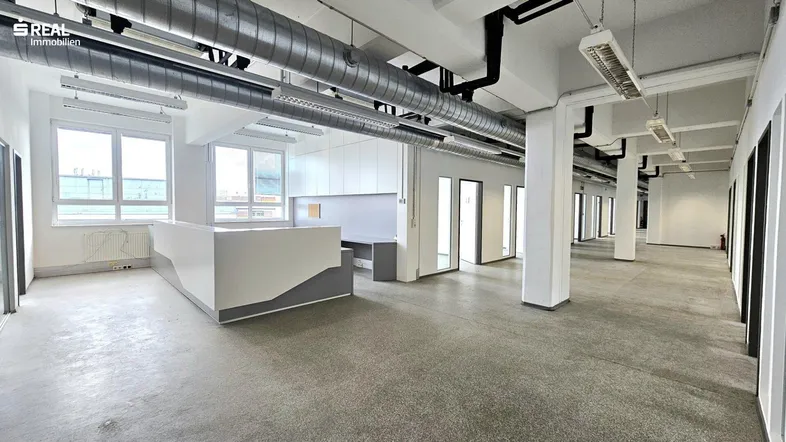 Großes, offenes Büro mit Betonboden, sichtbaren Lüftungsrohren und viel Tageslicht durch die Fenster.
