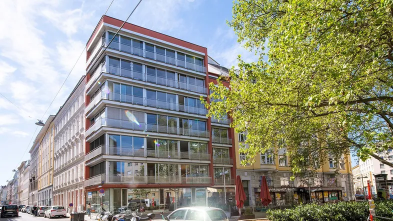 Modernes Eckgebäude mit vielen Glasflächen und Balkonen an einer belebten Stadtstraße unter blauem Himmel.