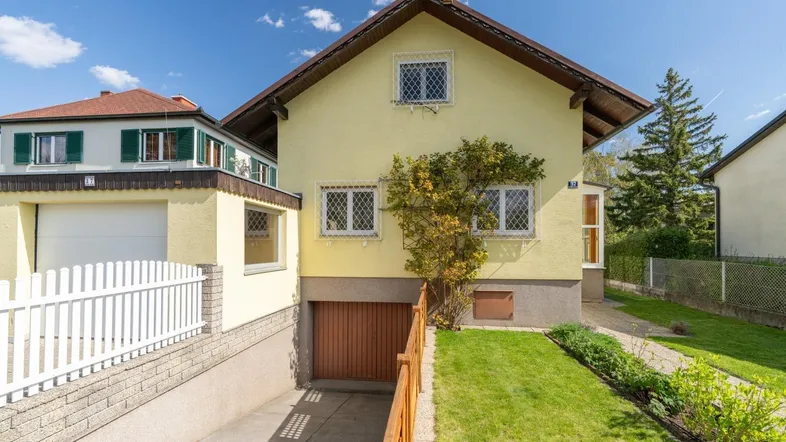 Gelbes Einfamilienhaus mit Satteldach, Vorgarten und Zufahrt zur Tiefgarage unter blauem Himmel.