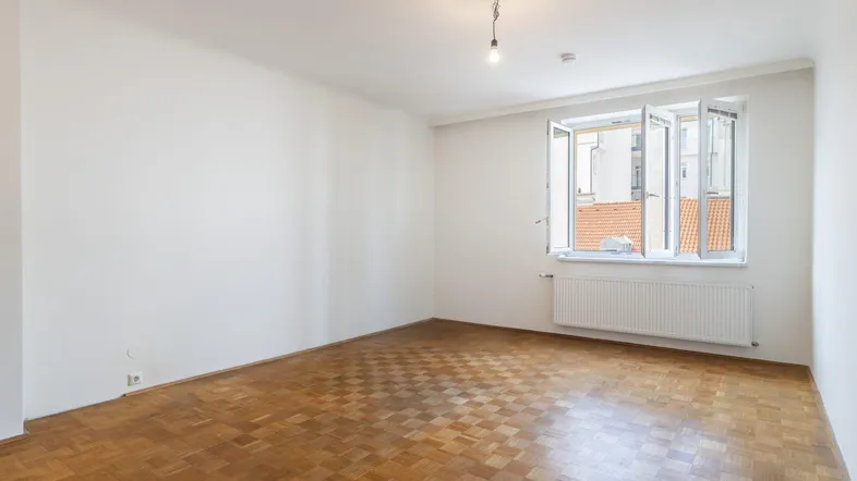 Helles, leeres Zimmer mit Parkettboden und einem Fenster, das einen Blick auf die umliegenden Gebäude bietet.