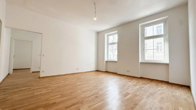 Helles, leeres Zimmer mit Parkettboden und zwei Fenstern, bereit zur individuellen Gestaltung.