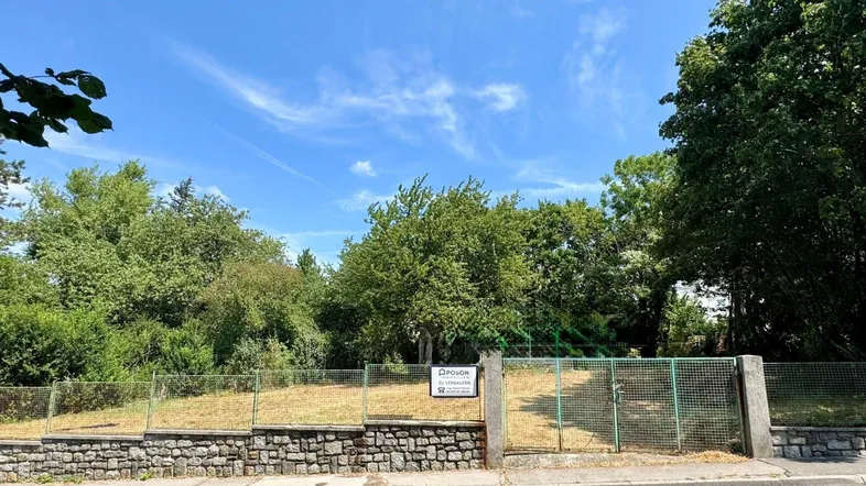 Unbebautes Grundstück mit grüner Umzäunung und Steinmauer, umgeben von üppiger Vegetation unter klarem blauem Himmel.