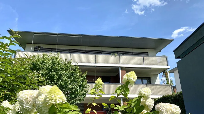 Modernes Mehrfamilienhaus mit mehreren Balkonen und üppiger Bepflanzung im Vordergrund.