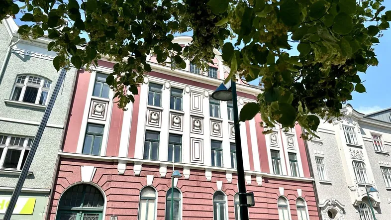 Historisches Gebäude mit rosa Fassade und dekorativen Elementen unter grünen Bäumen.