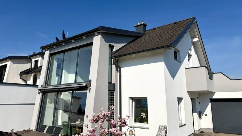 Moderne Hausfassade mit großen Fensterfronten und einer sonnigen Terrasse im Vordergrund.