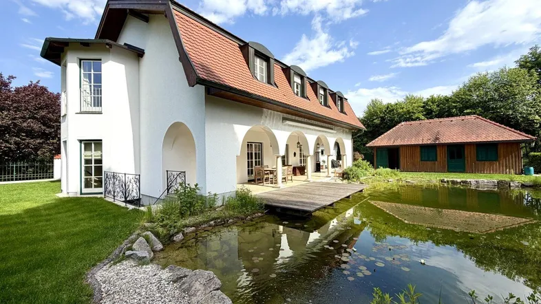 Großzügiges Anwesen mit weißer Fassade, Mansarddach und einem idyllischen Teich im Gartenbereich.