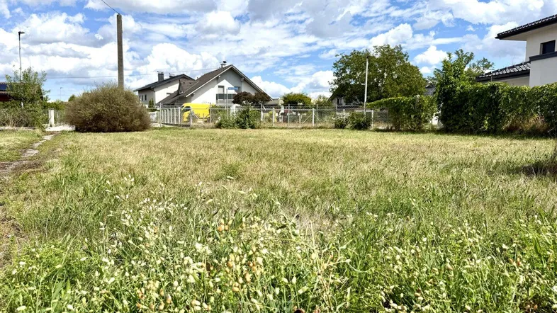 Großes, unbebautes Grundstück mit hohem Gras und umliegenden Wohnhäusern unter bewölktem Himmel.