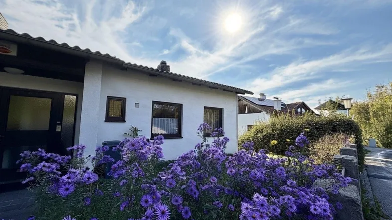 Sonnenbeschienenes Haus mit lila Blumen im Vorgarten und blauem Himmel.