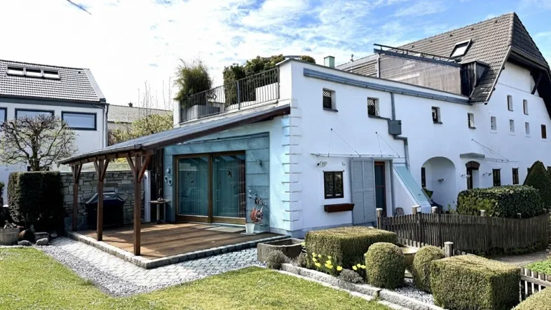 Weißes Haus mit Holzterrasse und Pergola, umgeben von einem gepflegten Garten unter blauem Himmel.
