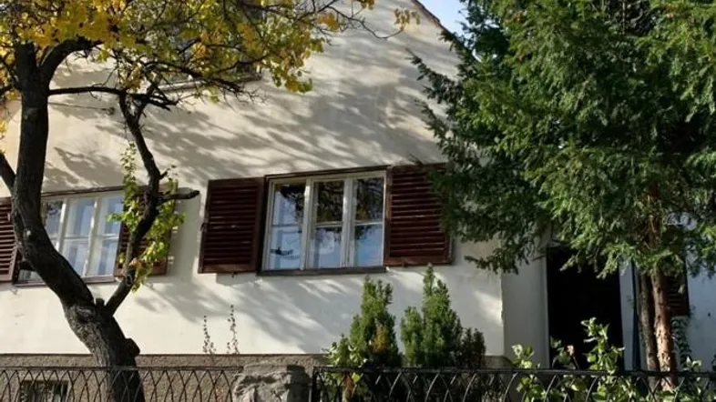 Fassade eines Hauses mit herbstlichen Bäumen und einer Steinmauer im Vordergrund, die das Grundstück begrenzt.