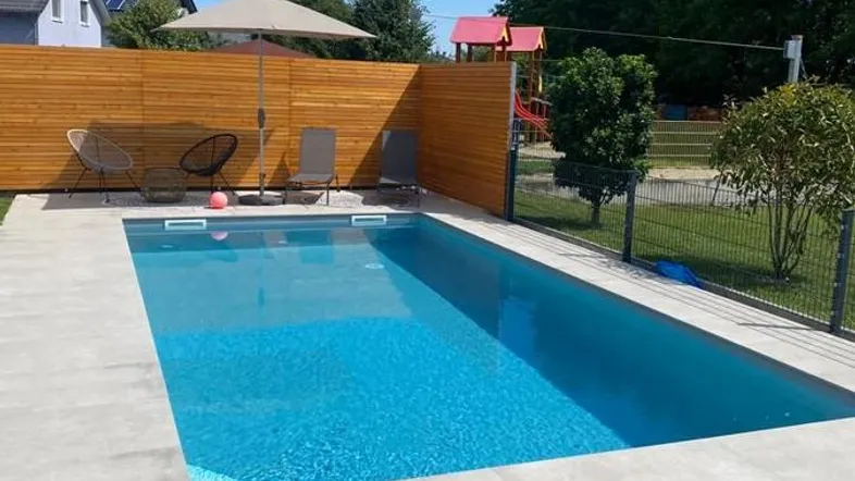 Moderner Außenbereich mit großem Pool, Liegeflächen und Holzumzäunung unter blauem Himmel.