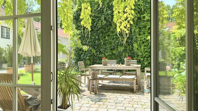Gemütliche Terrasse mit Holztisch und Stühlen, umgeben von üppigem Grün und Kletterpflanzen.