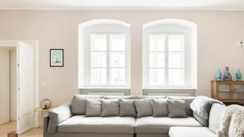 Helles Wohnzimmer mit beigefarbenen Wänden, großem Ecksofa und zwei hohen Bogenfenstern.