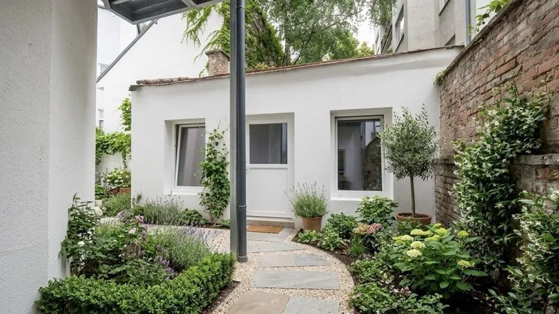 Kleiner, gepflegter Garten mit Steinweg, üppiger Bepflanzung und Zugang zu einem weißen Gebäude.
