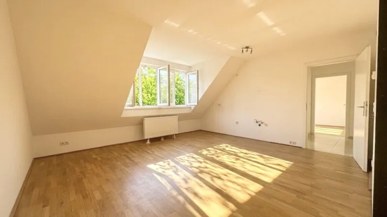 Helles Zimmer im Dachgeschoss mit Holzboden und schrägen Wänden, durchflutet von Sonnenlicht.